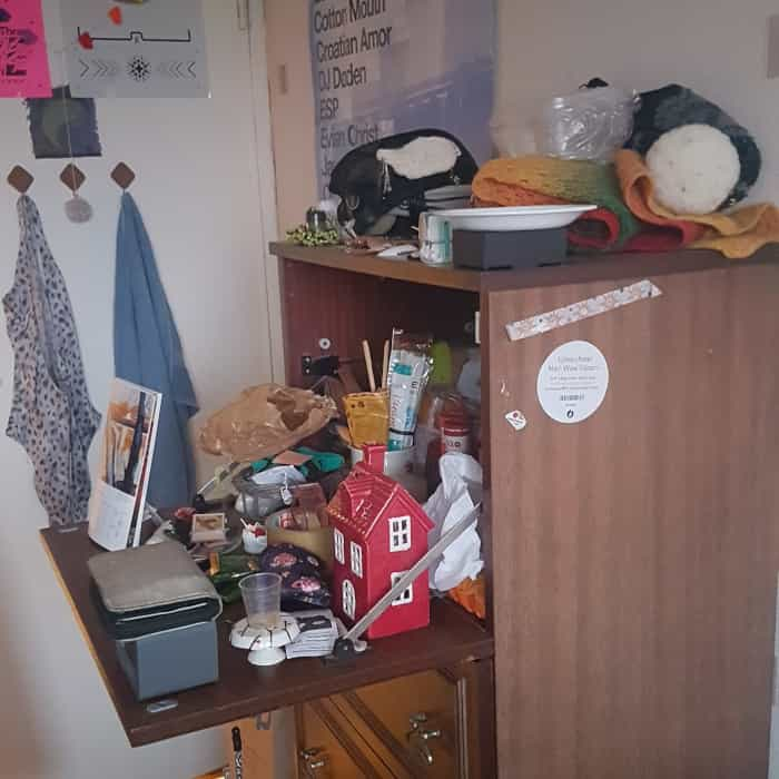 a tall chest of drawers, the top part with a door opening downward all shelflike, with many things cluttered on top of it