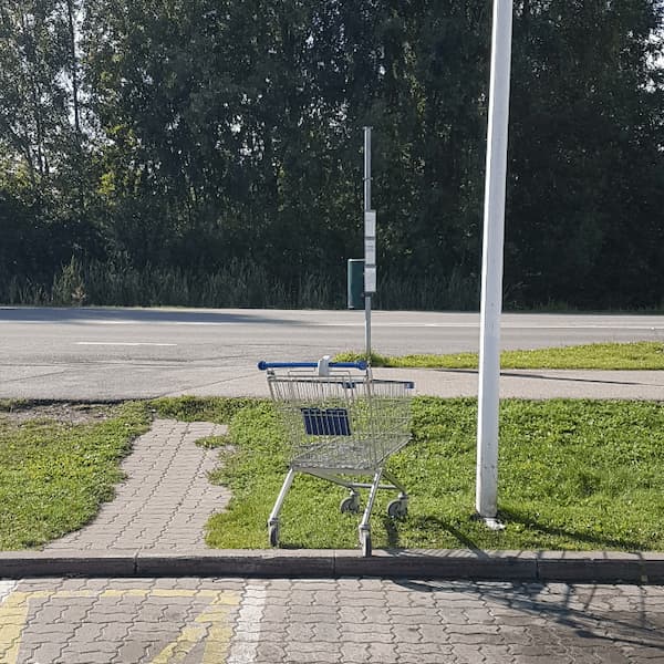 a shopping cart stood next to a flagpost at the edge of a parking lot, near a bus stop