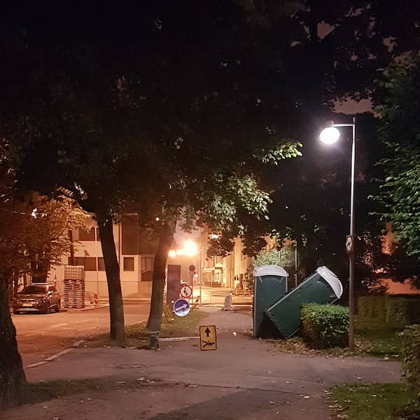 tree-lined sidewalk with a detour sign fallen across, and two portapotties, one tilted back against a hedge