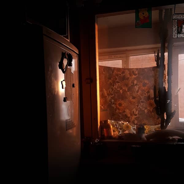 dim kitchen corner, lit only by sunset, showing the fridge and the window to the inward balcony