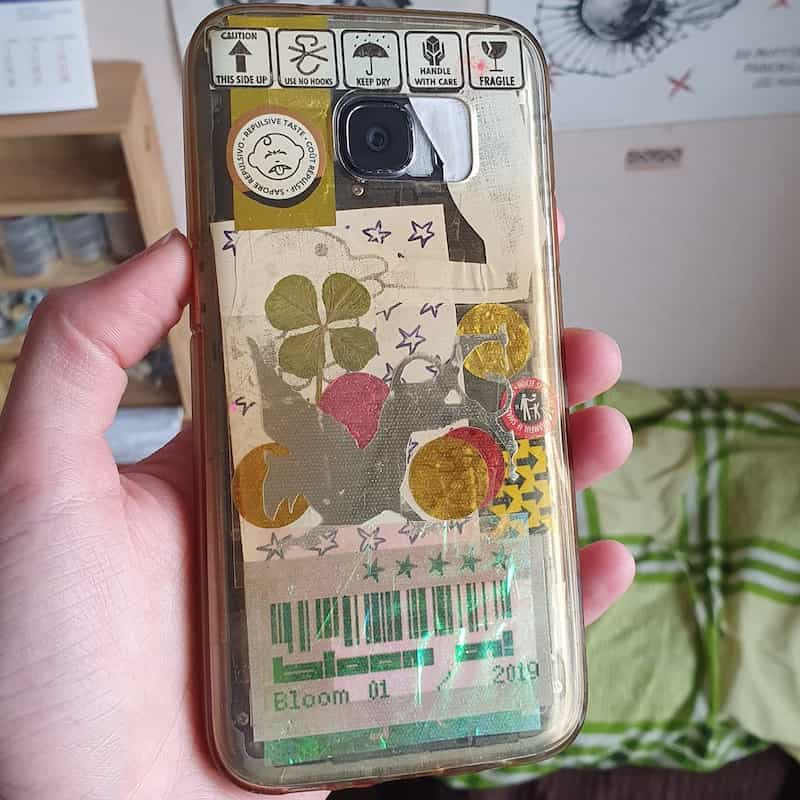 the back of the phone, with the inside of its transparent case stacked with various art, warning symbols, confetti pieces, a 4-leaf clover, silver dragon, 'bloom 01!' with barcode, etc