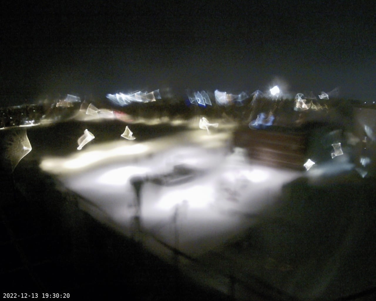 a view from the camera on december 13 2022, 7:30pm, a parking lot and some buildings seen. the street lights distorted into fun shapes from the snowmelt on the lens